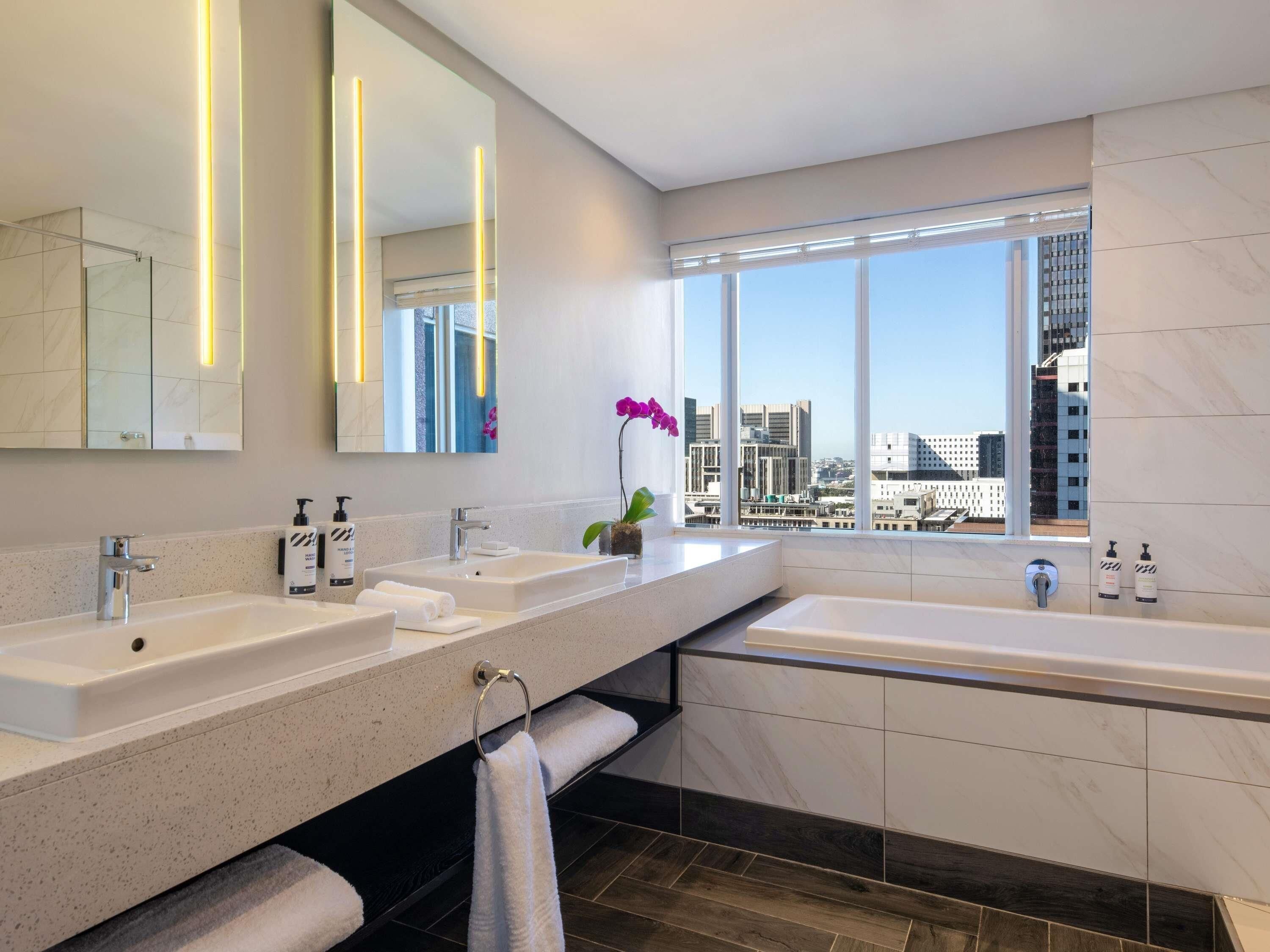 Een moderne badkamer met dubbele wastafels, een grote spiegel, een badkuip en uitzicht op de stad door grote ramen.