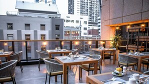 Un área de comedor en la azotea con mesas y sillas, una planta en maceta y vistas de la ciudad.