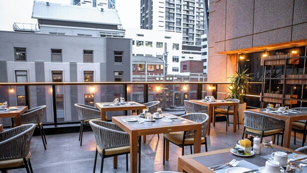 Un área de comedor en la azotea con mesas y sillas, una planta en maceta y vistas de la ciudad.