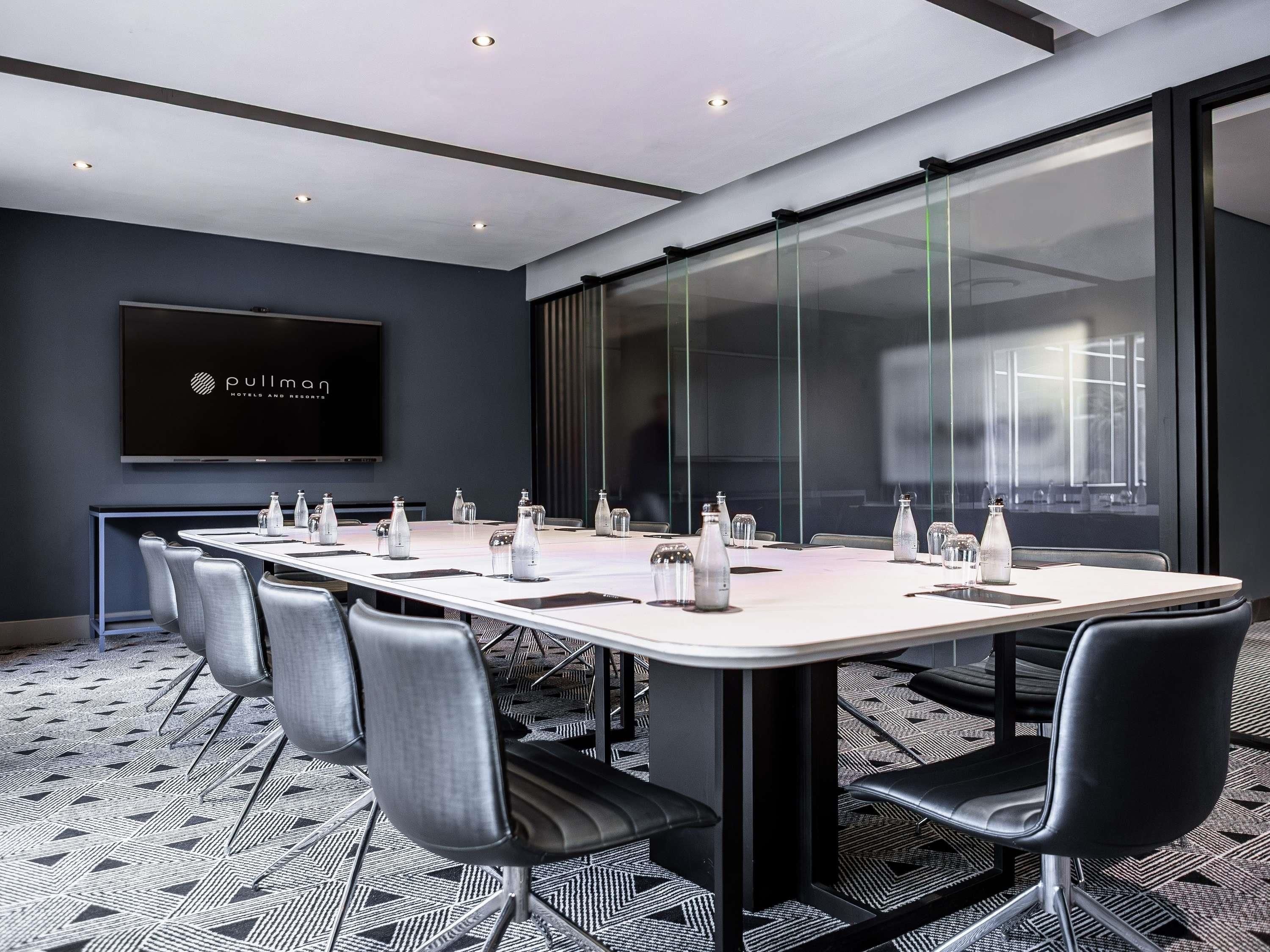 A conference room with a long table, chairs, a TV screen, and a glass wall.