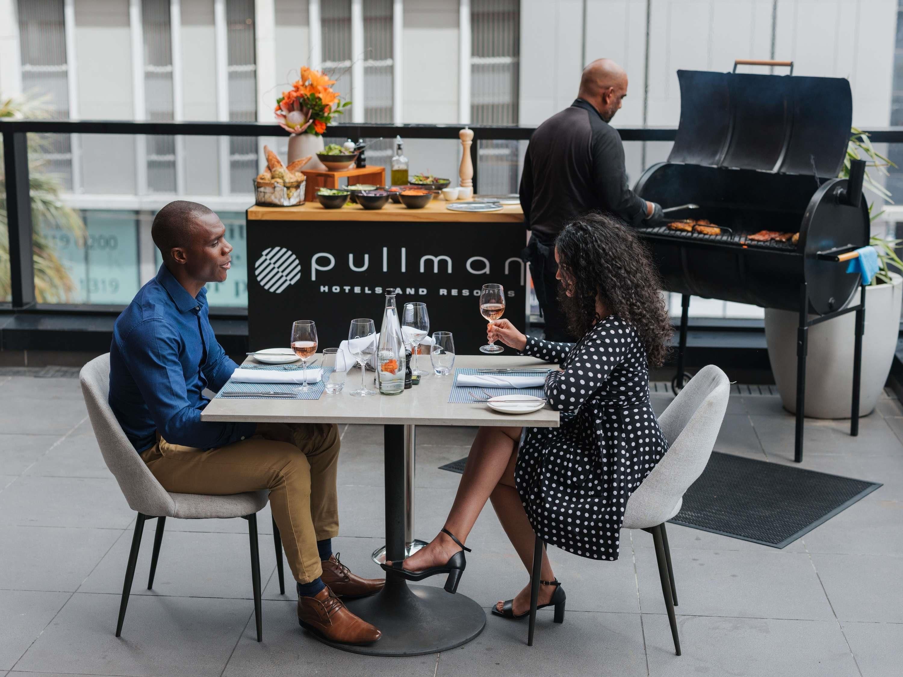 A Pullman Hotels and Resorts outdoor dining area with a grill, tables set with plates, glasses, and a vase of flowers.