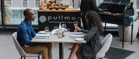 A Pullman Hotels and Resorts outdoor dining area with a grill, tables set with plates, glasses, and a vase of flowers.