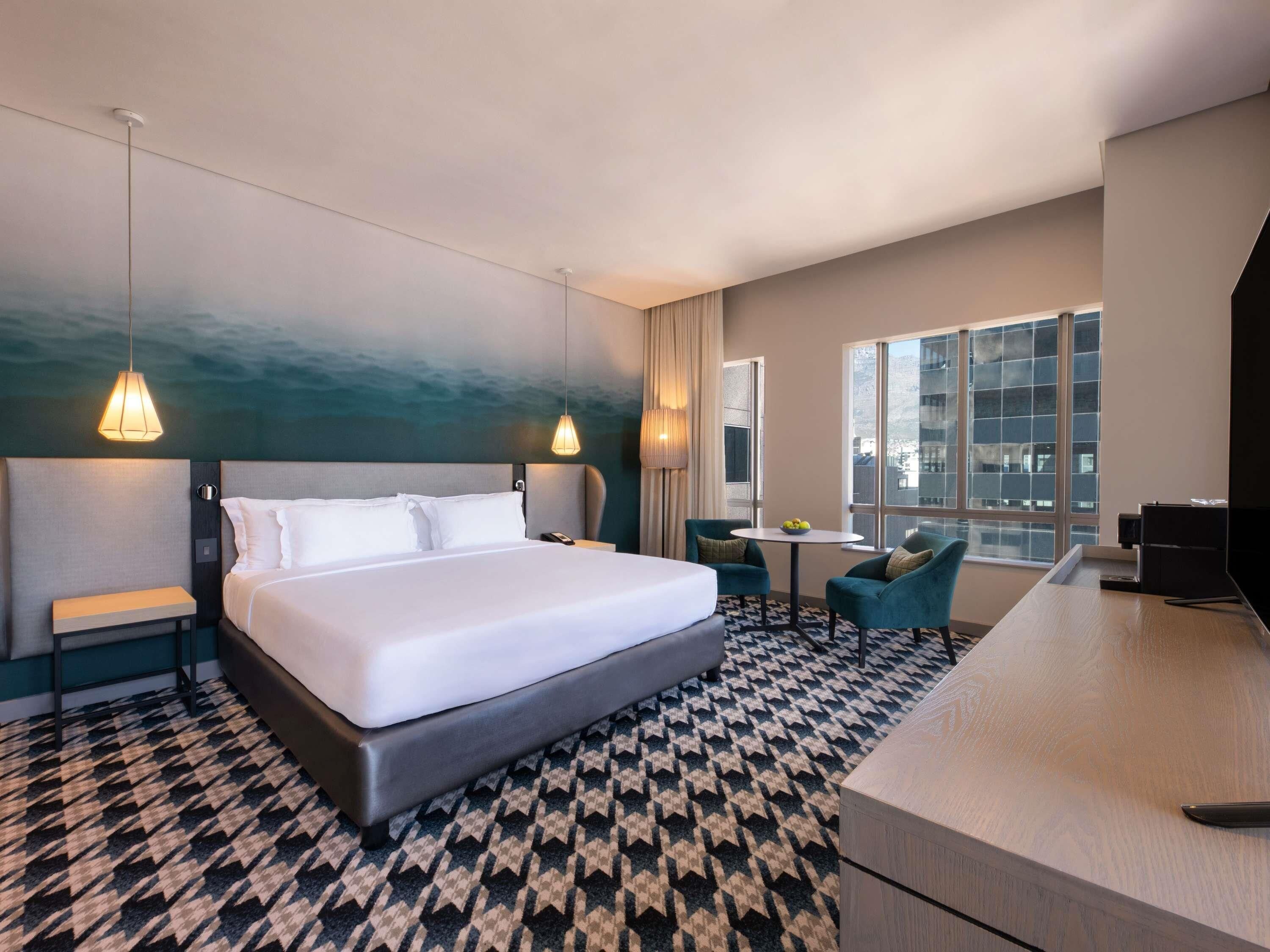 Habitación de hotel con una cama grande, mesitas de noche, un escritorio y una silla. Hay una ventana con vistas a edificios.