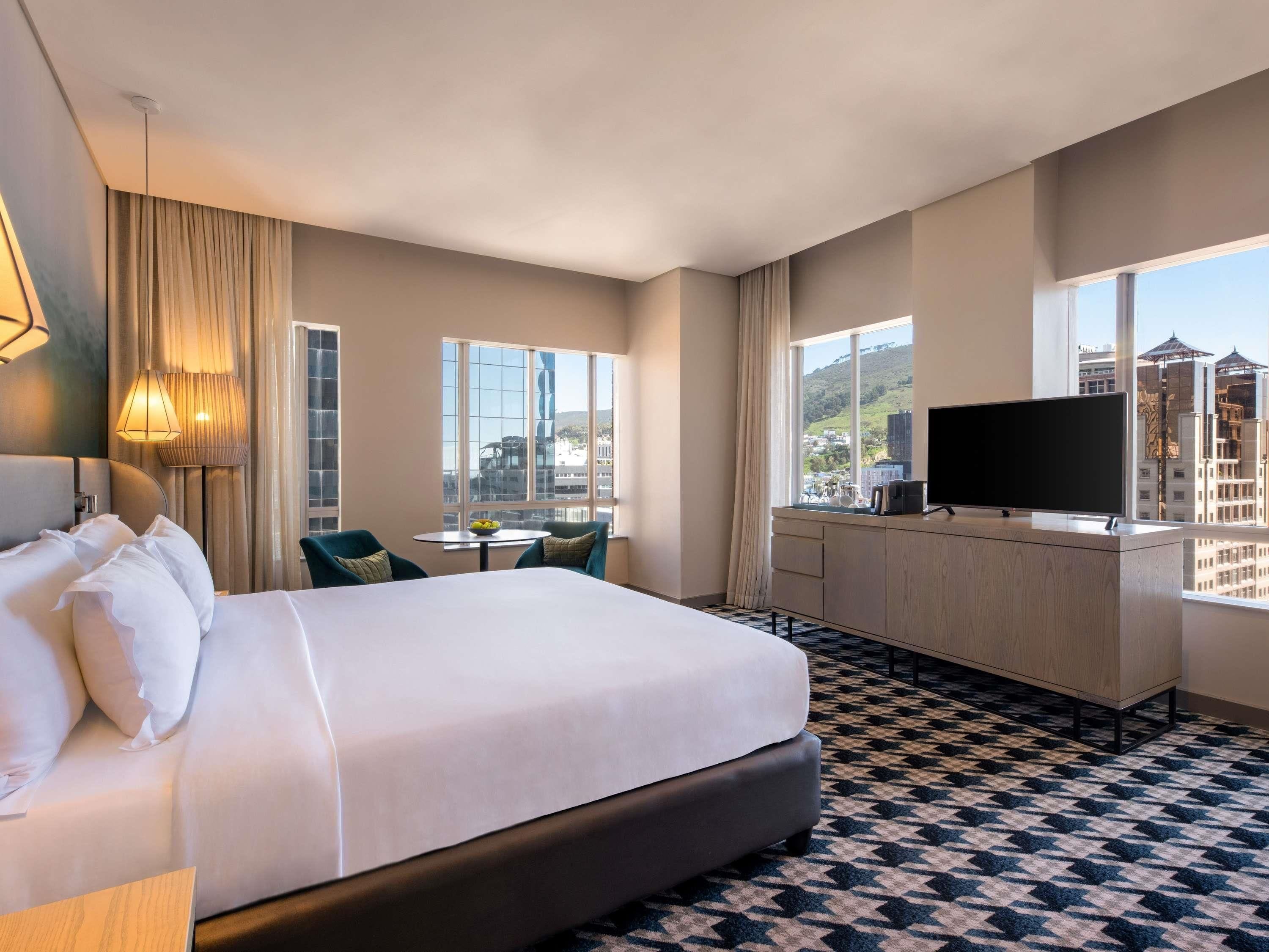 A hotel room with a large bed, a desk with a TV, a small table with chairs, and large windows offering a city view.