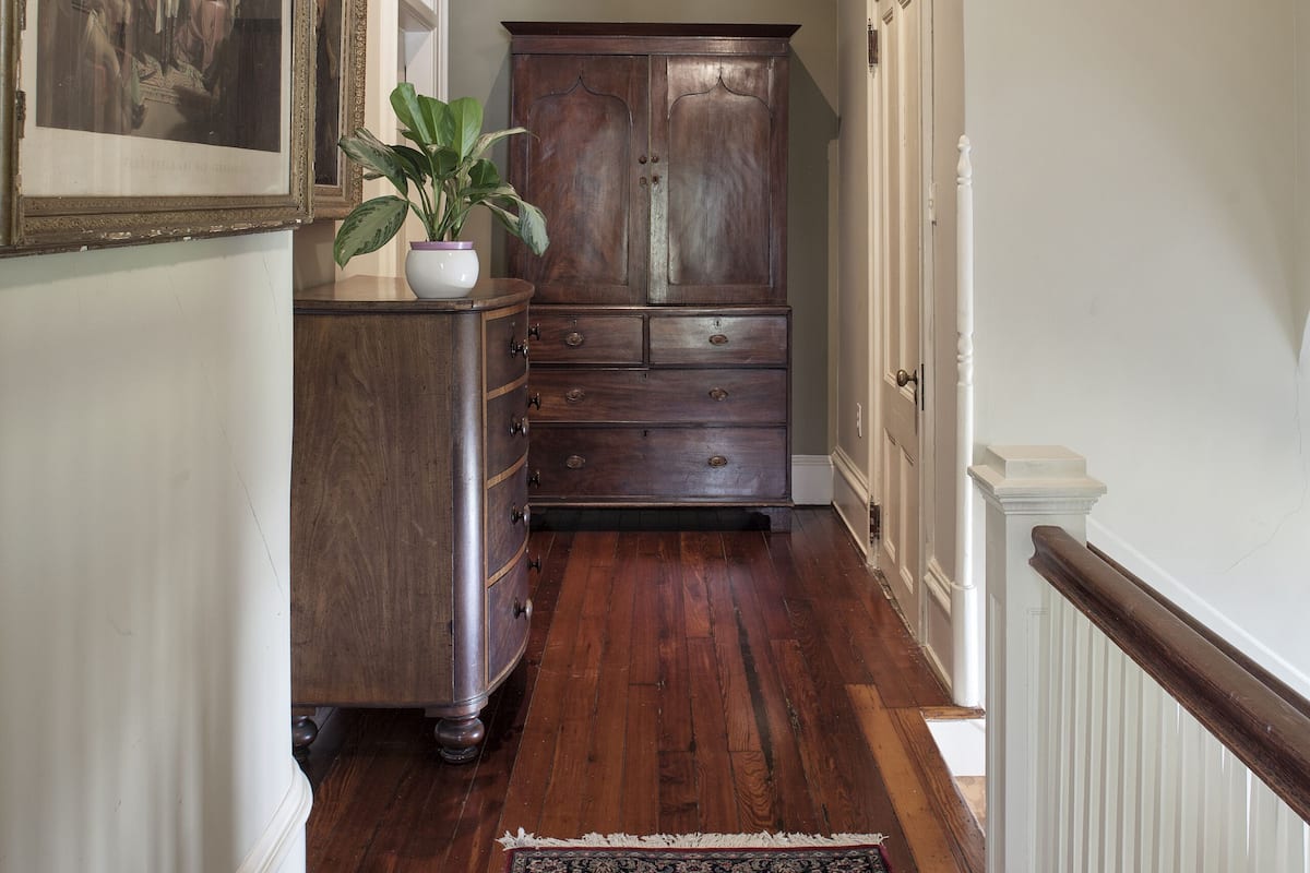 A hallway with wooden flooring, a large dark wooden cabinet, a potted plant, and a framed painting on the wall.