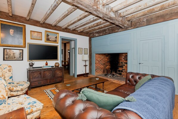 A traditional living room with exposed wooden beams, a fireplace, a leather sofa, a wooden cabinet, and a flat-screen TV.
