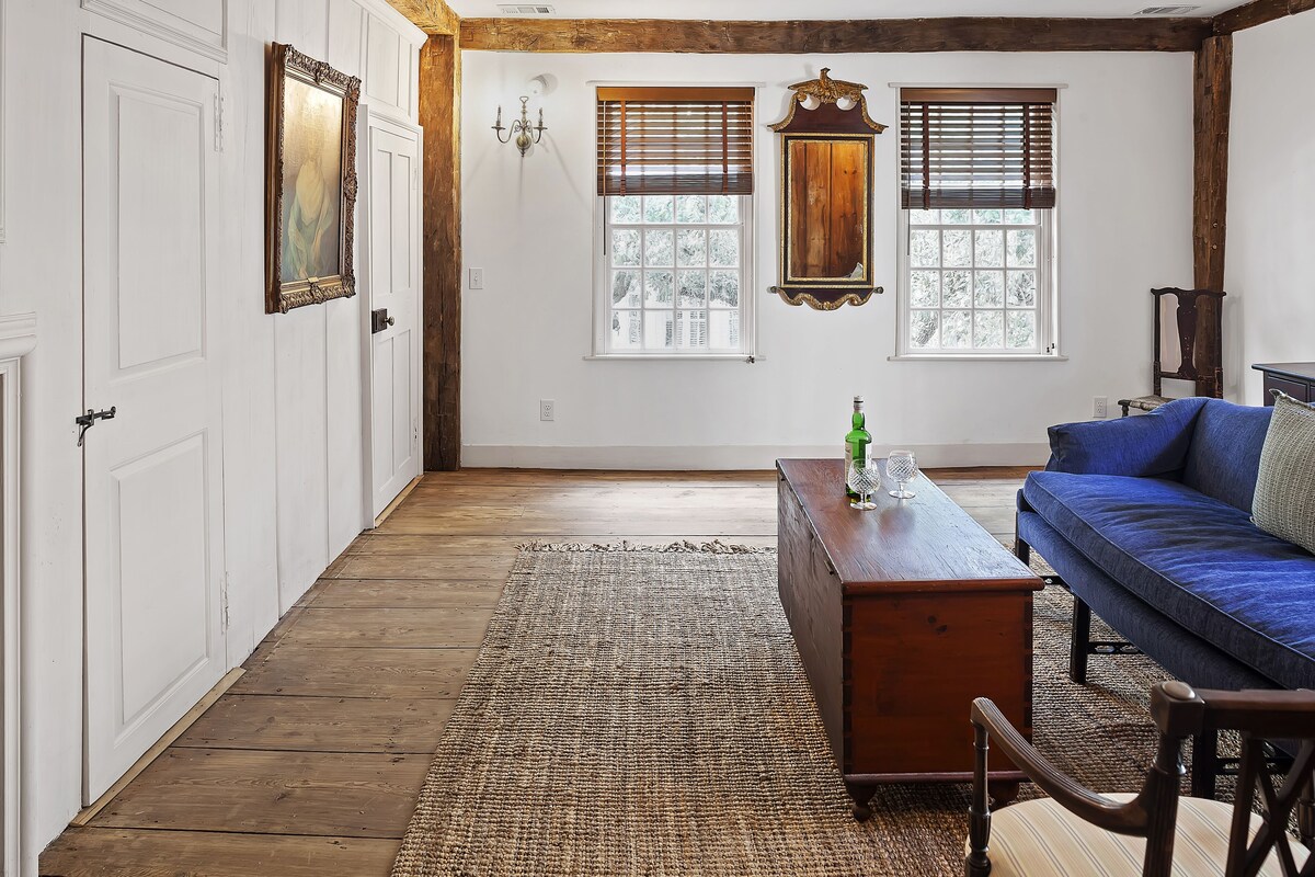 A room with a wooden floor, a blue sofa, a wooden table, and two windows with blinds.