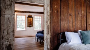 A rustic room with wooden floors, a bed with white and green pillows, and two windows with a view of greenery.
