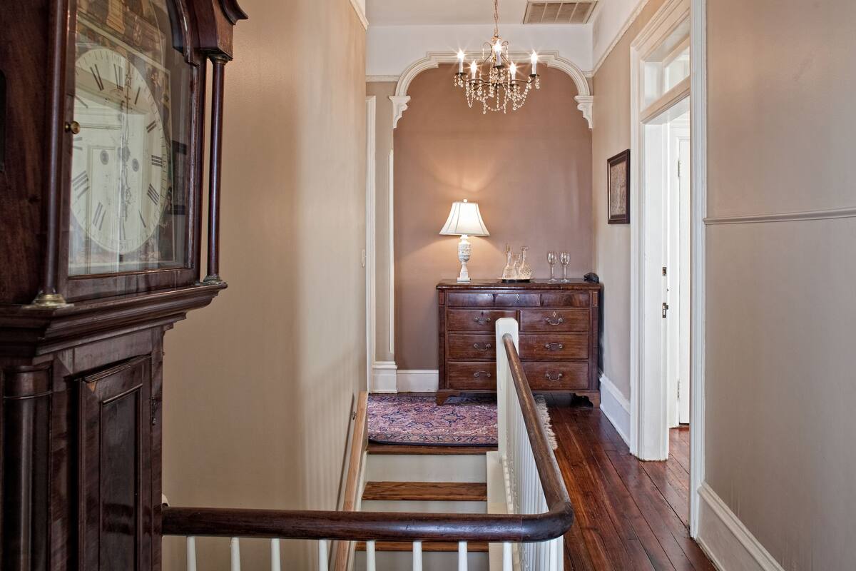 A hallway with a wooden staircase, a chandelier, a wooden dresser, and a large clock.