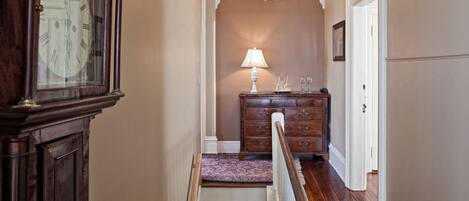 A hallway with a wooden staircase, a chandelier, a wooden dresser, and a large clock.