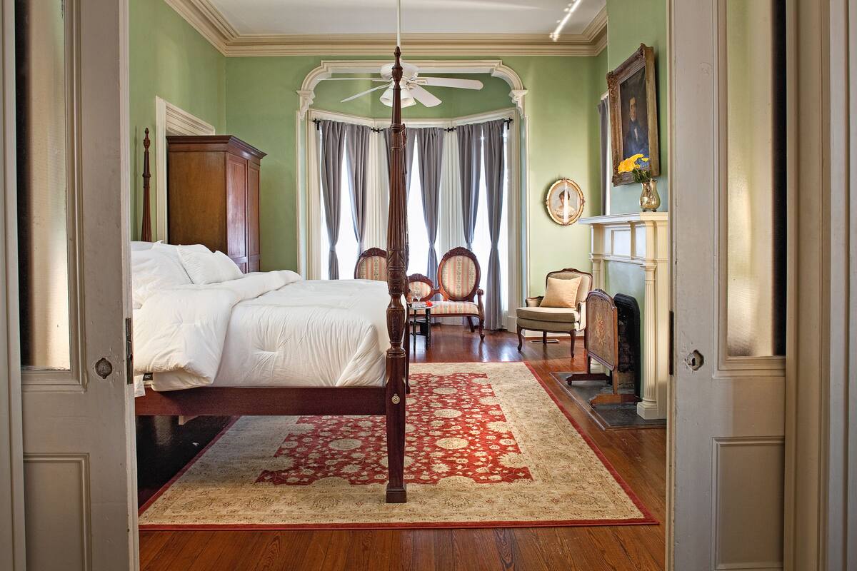A bedroom with a four-poster bed, a wooden floor, a red and beige patterned rug, a ceiling fan, and a fireplace with a vase of flowers.