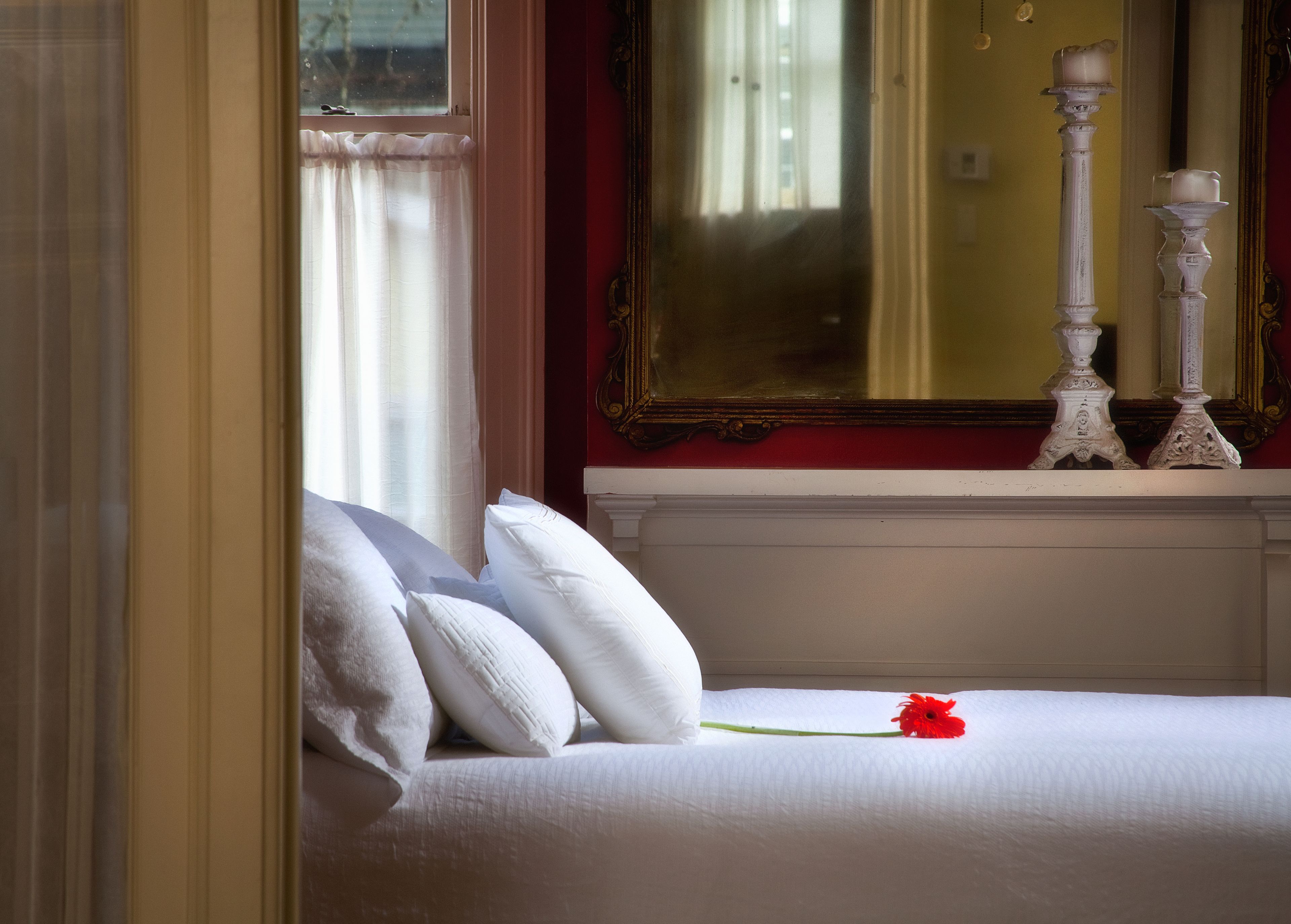 A bedroom with a bed, white pillows, a single red flower, and a mirror above the bed.
