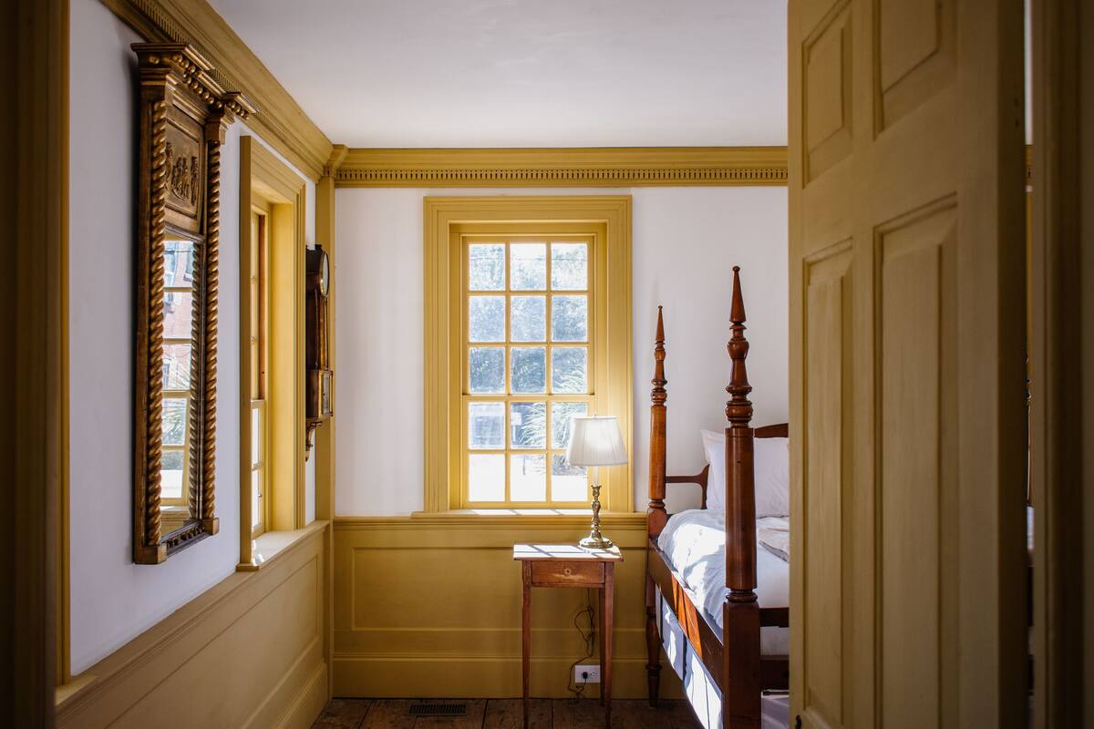 A bedroom with a large window, a bedside table with a lamp, and a wooden bed.