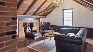 A loft-style room with exposed wooden beams, a brick wall, and a large window. There are two leather armchairs, a leather sofa, and a round coffee table.