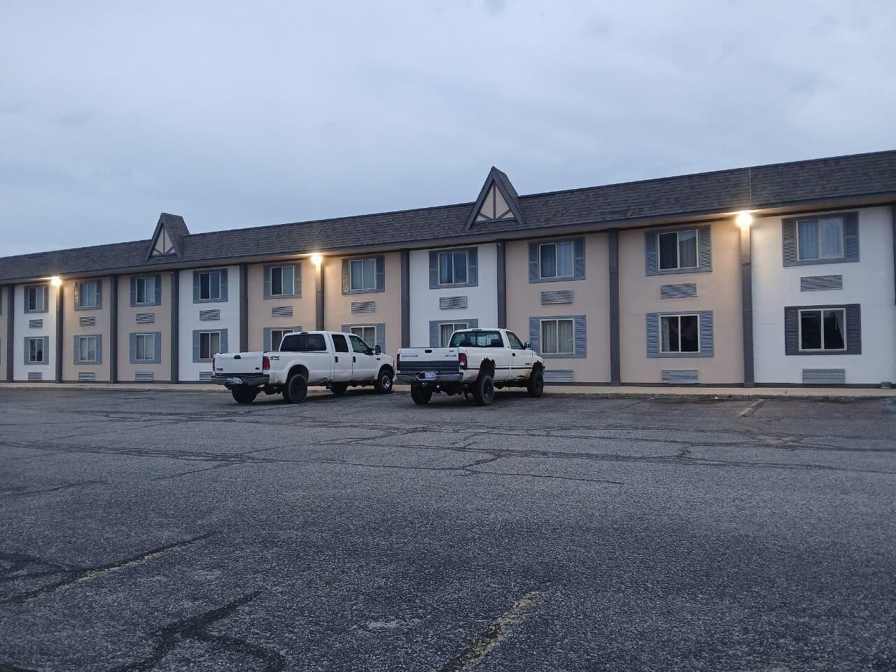 A motel with a row of rooms, each with a window and a door. There are two white pickup trucks parked in front.