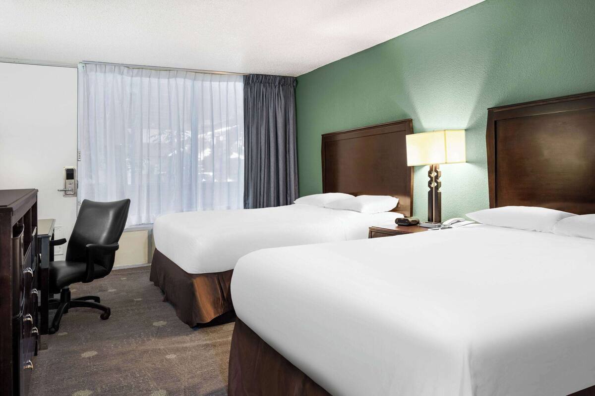 A hotel room with two beds, a desk, a chair, and a lamp.