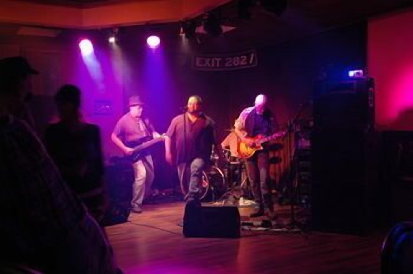 Una banda tocando en un escenario, con un letrero que dice 'EXIT 2827'.
