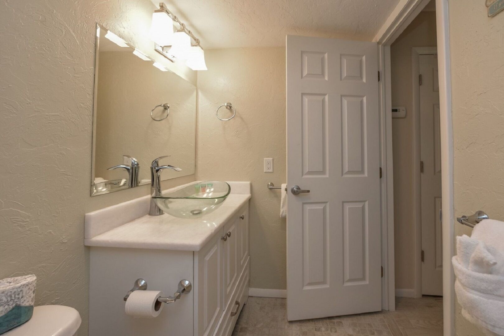 Un baño con lavamanos de vidrio, dos grifos, un inodoro y una puerta.