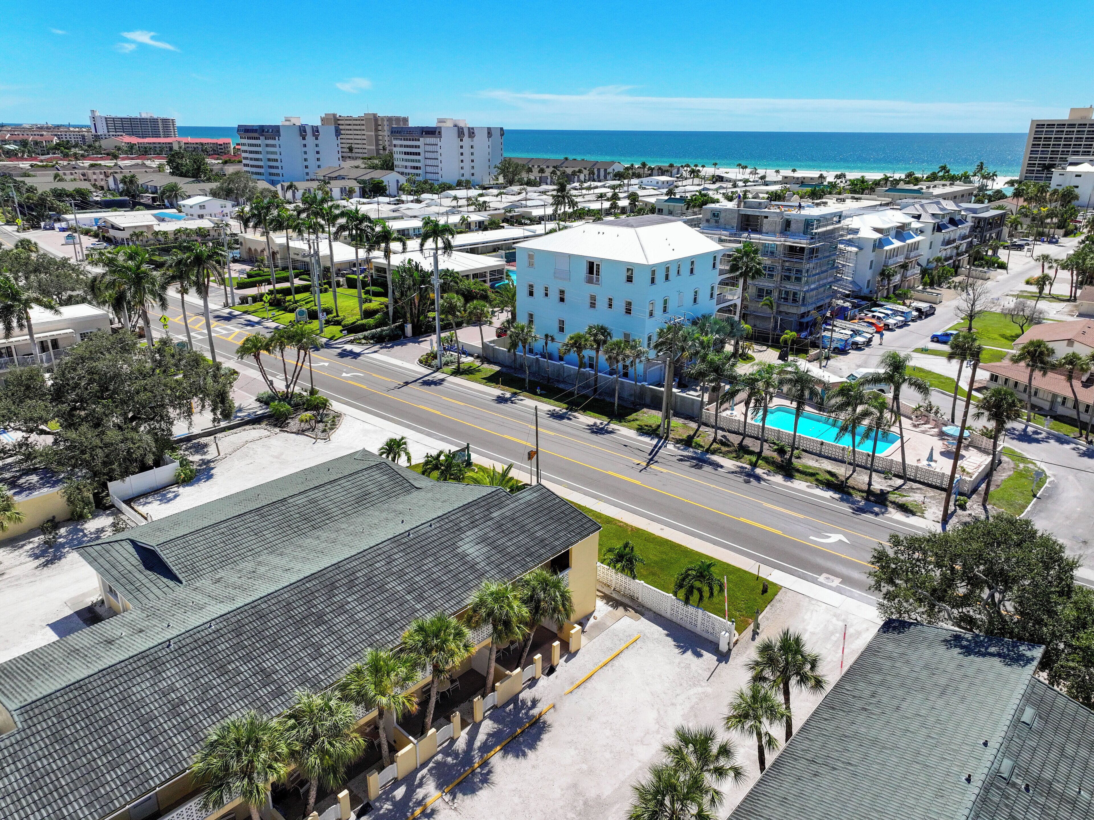 A coastal hotel with multiple buildings, a swimming pool, and a road with palm trees.