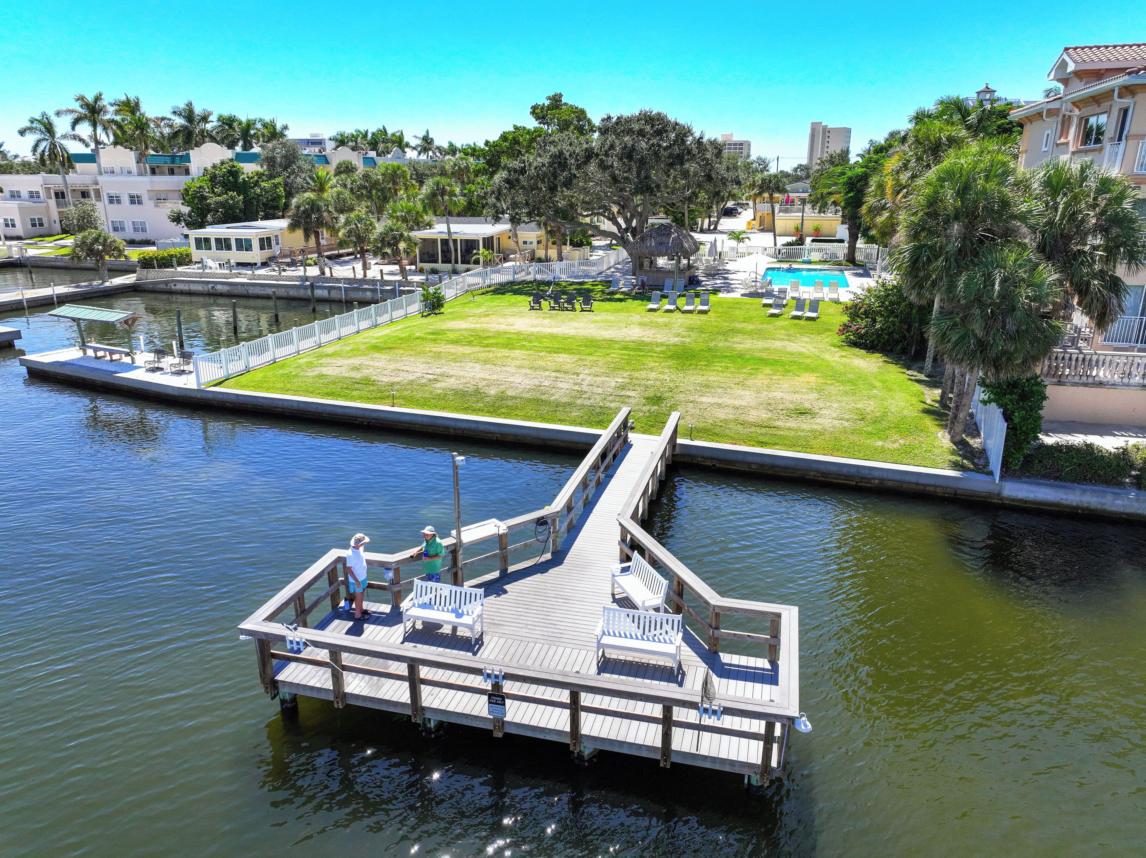 A dock with a bench and a small boat, a grassy area with a pool, and buildings in the background.