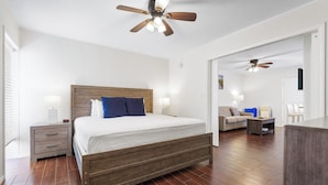 A bedroom with a wooden bed, a nightstand, a ceiling fan, and a view into another room with a sofa and chairs. - Sea Club II by Beachside Management (Siesta Key)