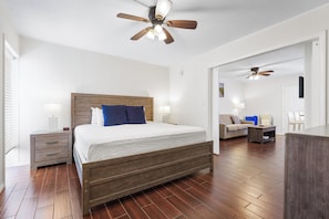 Un dormitorio con cama de madera, una mesita de noche, un ventilador de techo y vista a otra habitación con un sofá y sillones.