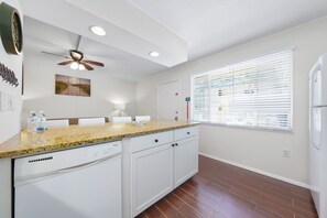 A kitchen with a granite countertop, white cabinets, a dishwasher, a ceiling fan, and a window with blinds. - Sea Club II by Beachside Management (Siesta Key)
