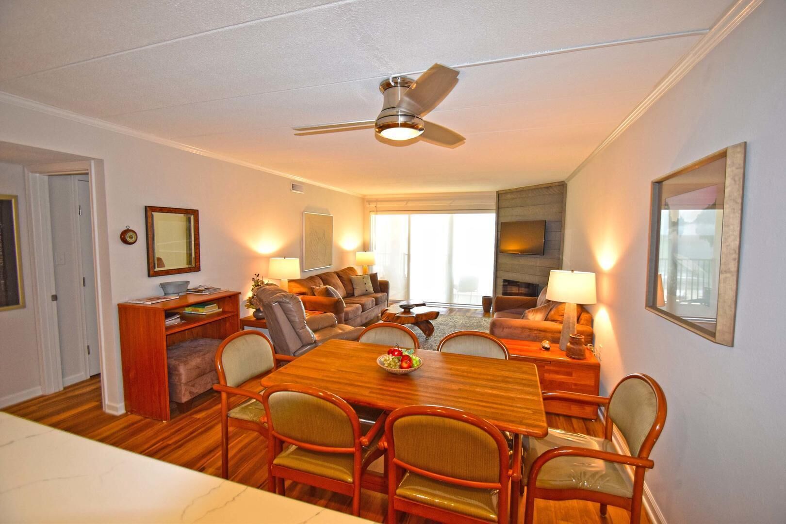 A spacious living room with a wooden dining table, comfortable seating, a ceiling fan, and a large window providing natural light.
