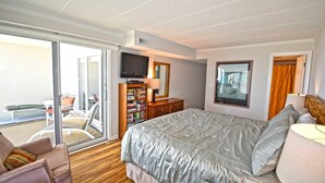 A hotel room with a large bed, a TV, a bookshelf, a desk, and a balcony with outdoor seating.