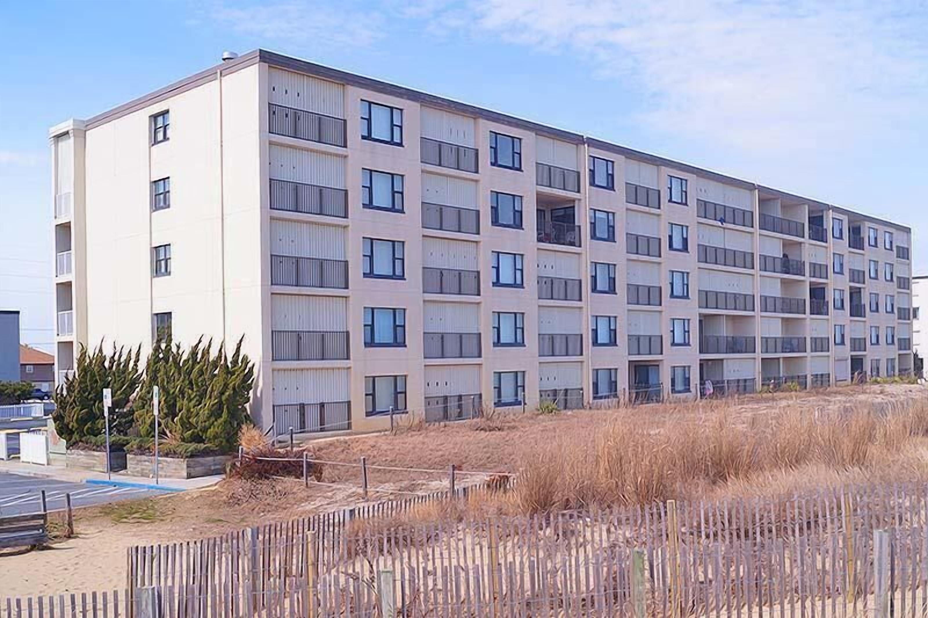A multi-story residential building with a flat facade, balconies, and a parking area in front.