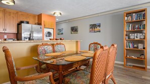 A compact kitchen with wooden cabinets, a stainless steel refrigerator, a built-in oven, and a washing machine. There is a dining area with a glass table and wicker chairs, and a bookshelf filled with books and decorative items.