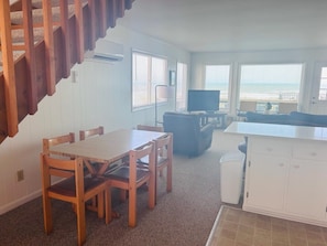 A living room with a wooden dining table, chairs, a sofa, a TV, and a kitchen area with cabinets.