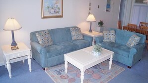 Un salon avec deux canapés bleus, une table basse blanche et deux tables d’appoint avec des lampes.