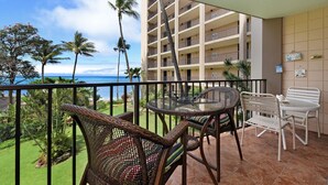 A balcony with wicker chairs and a table, overlooking a beach and a multi-story building with balconies. - Valley Isle 302 by RedAwning (Lahaina)