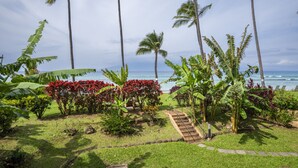 A tropical garden with palm trees and red flowers, a pathway, and a view of the ocean. - Paki Maui #109 1 Bedroom Condo by RedAwning (Lahaina)