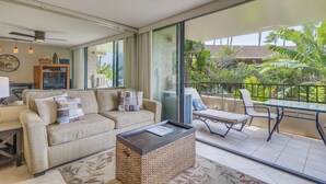 A spacious living area with a sofa, a coffee table, and a dining table with chairs. There is a balcony with a view of greenery and a building in the background. - Paki Maui #109 1 Bedroom Condo by RedAwning (Lahaina)