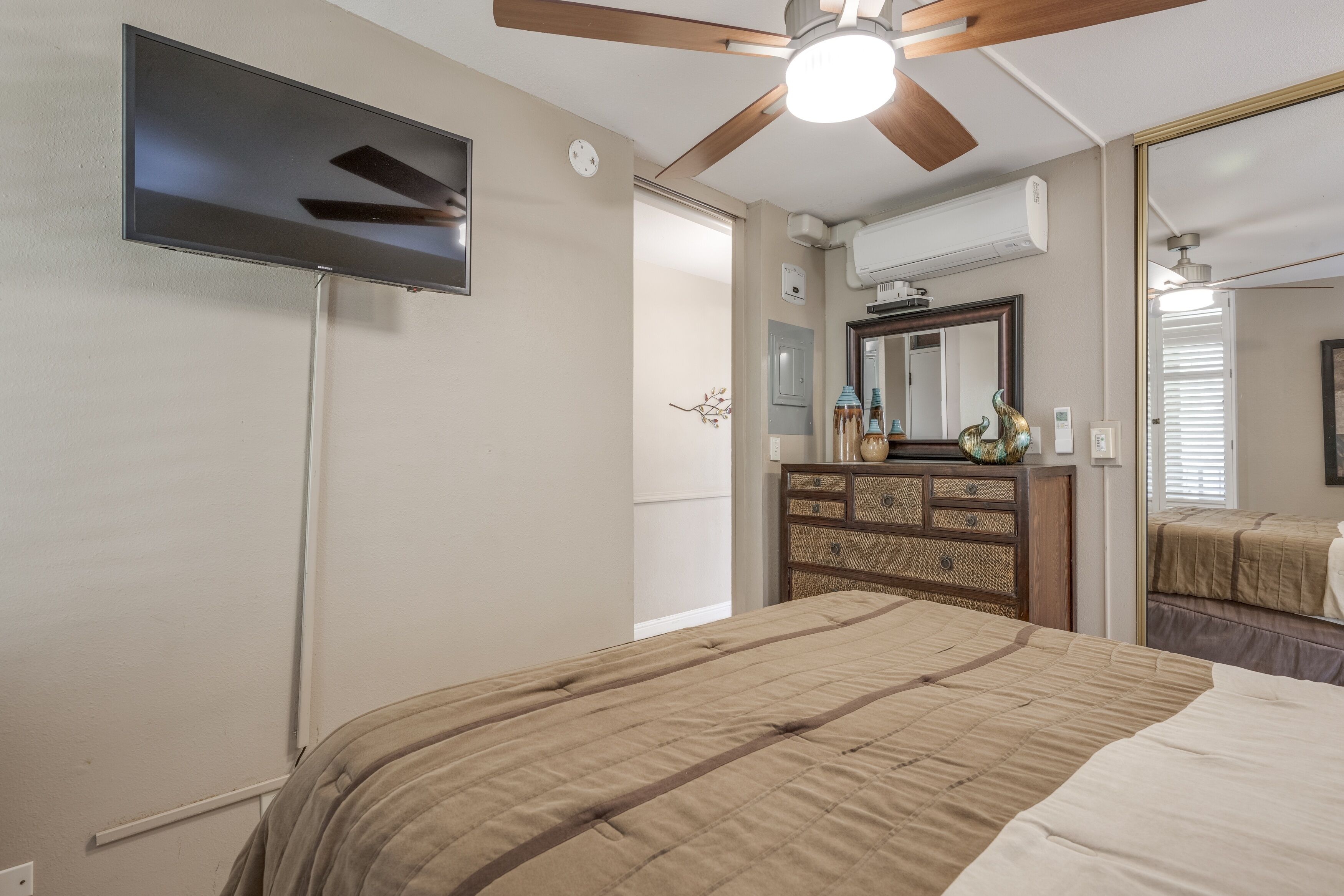 Ein Schlafzimmer mit einem Bett, einem Schminktisch, einem Spiegel, einem Deckenventilator und einem an der Wand montierten Fernseher.