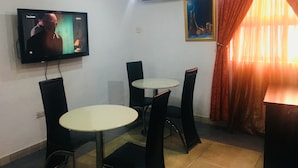 Una habitación con televisión montada en la pared, dos mesas redondas con sillas, un equipo de aire acondicionado, un cuadro en la pared y una ventana con cortinas.