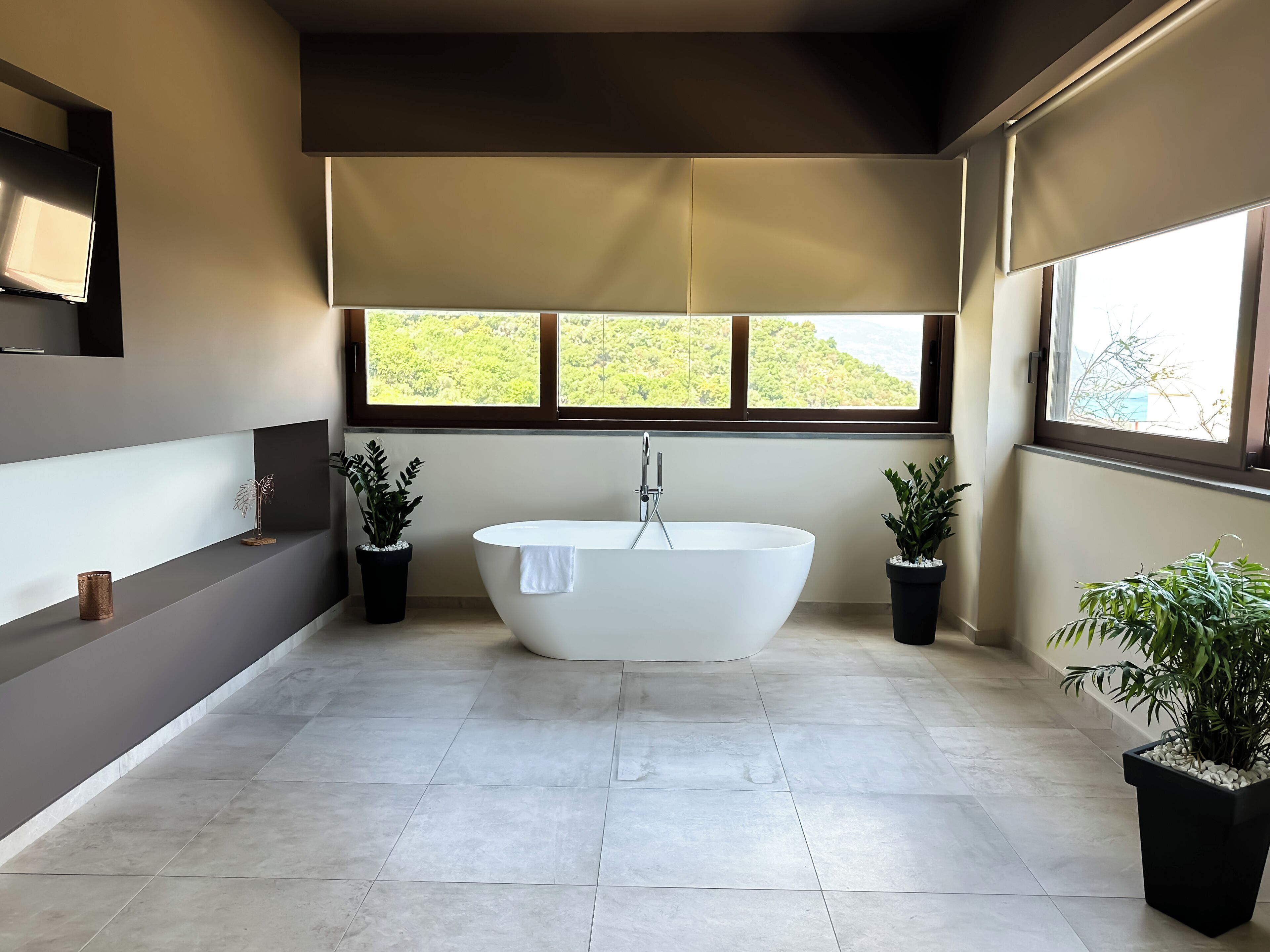 Un baño moderno con una bañera independiente, dos plantas en macetas y un ventanal con vistas a zonas verdes.