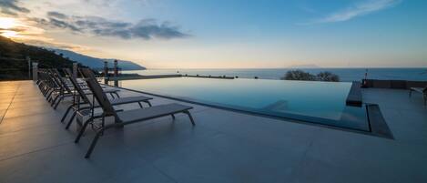 Una piscina infinita moderna con sillones de descanso, con vistas a un paisaje costero al atardecer.
