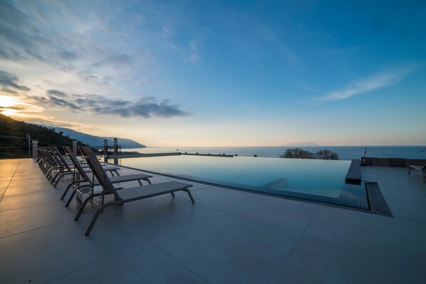 Una piscina infinita moderna con sillones de descanso, con vistas a un paisaje costero al atardecer.
