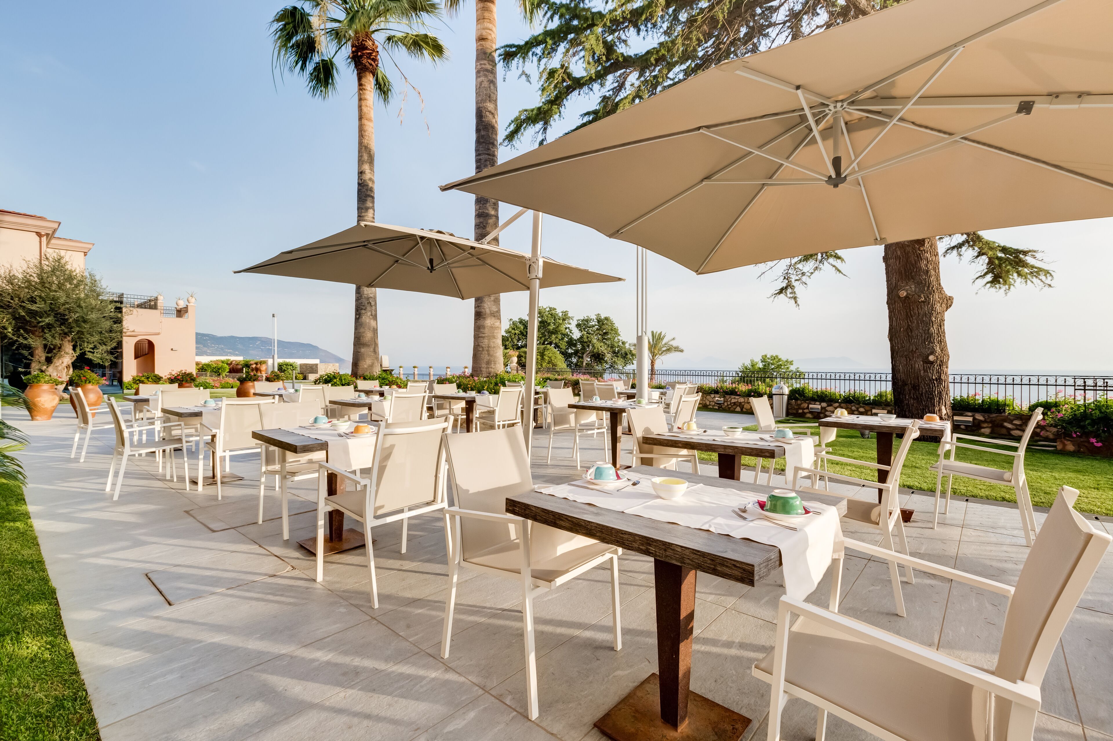 Un'area pranzo all'aperto con tavoli e sedie, grandi ombrelloni e vista sul mare.