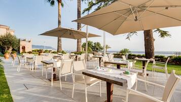 Un'area pranzo all'aperto con tavoli e sedie, grandi ombrelloni e vista sul mare.