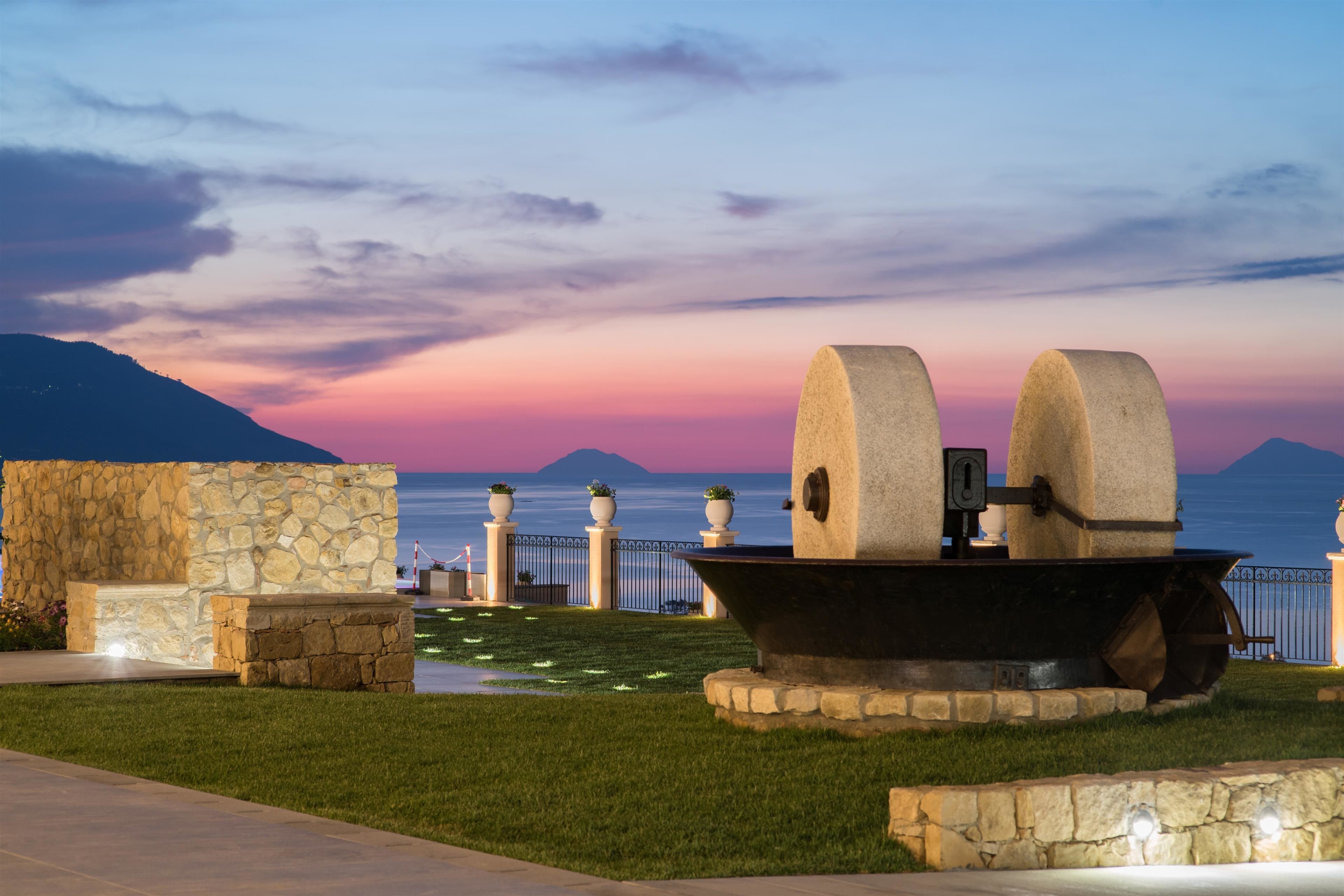 Una struttura in pietra con un'ampia vasca in primo piano, un muro di pietra e vista sul mare al tramonto.