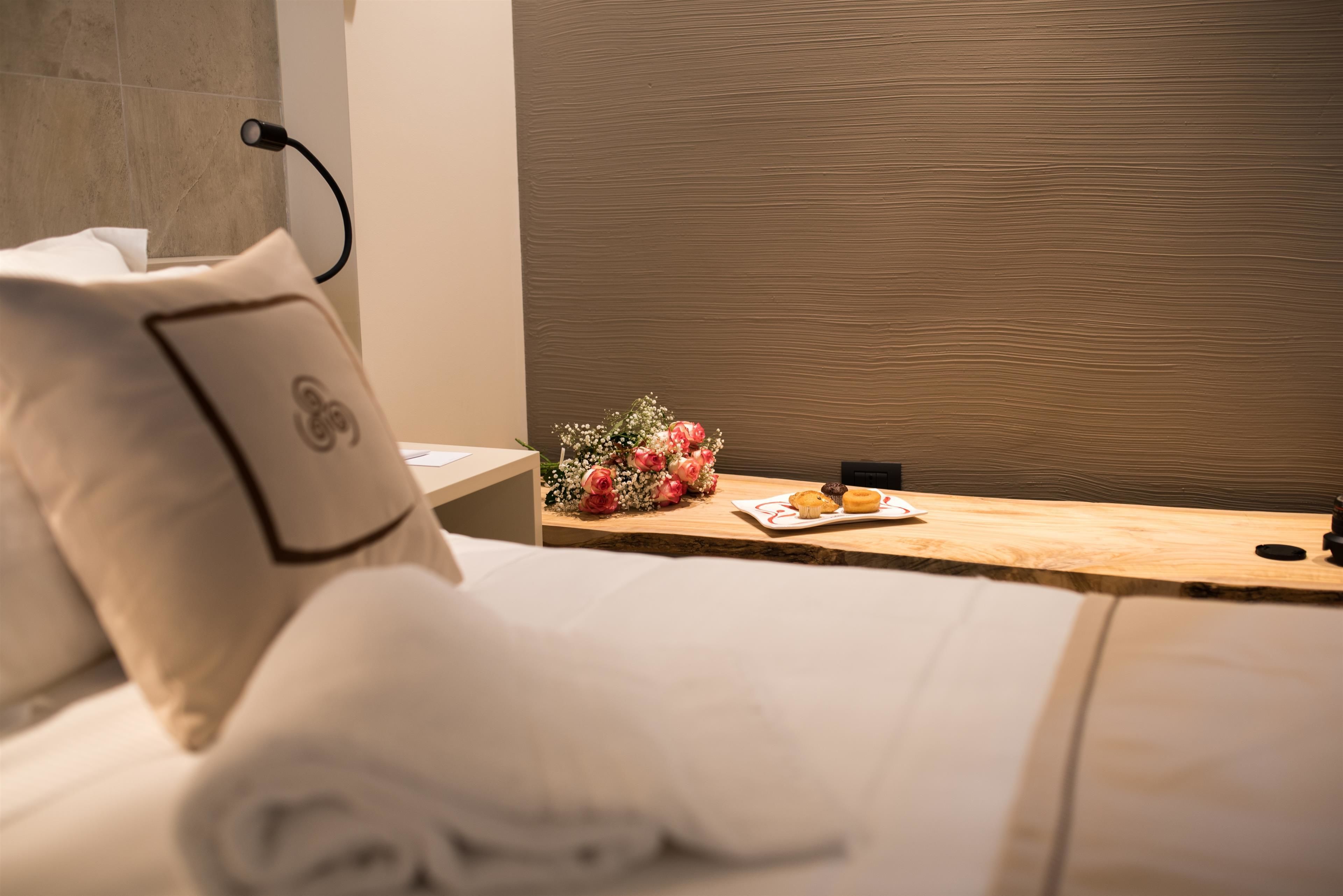 A neatly made bed with a pillow, a wooden bedside table with a bouquet of flowers and a plate of cookies, and a wall with a textured pattern.