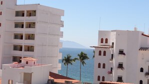 A coastal building with multiple balconies, a clear view of the sea, and palm trees in the foreground.