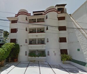 Un edificio de varios pisos con fachada blanca y detalles rojos, que incluye balcones y una entrada protegida por una reja.