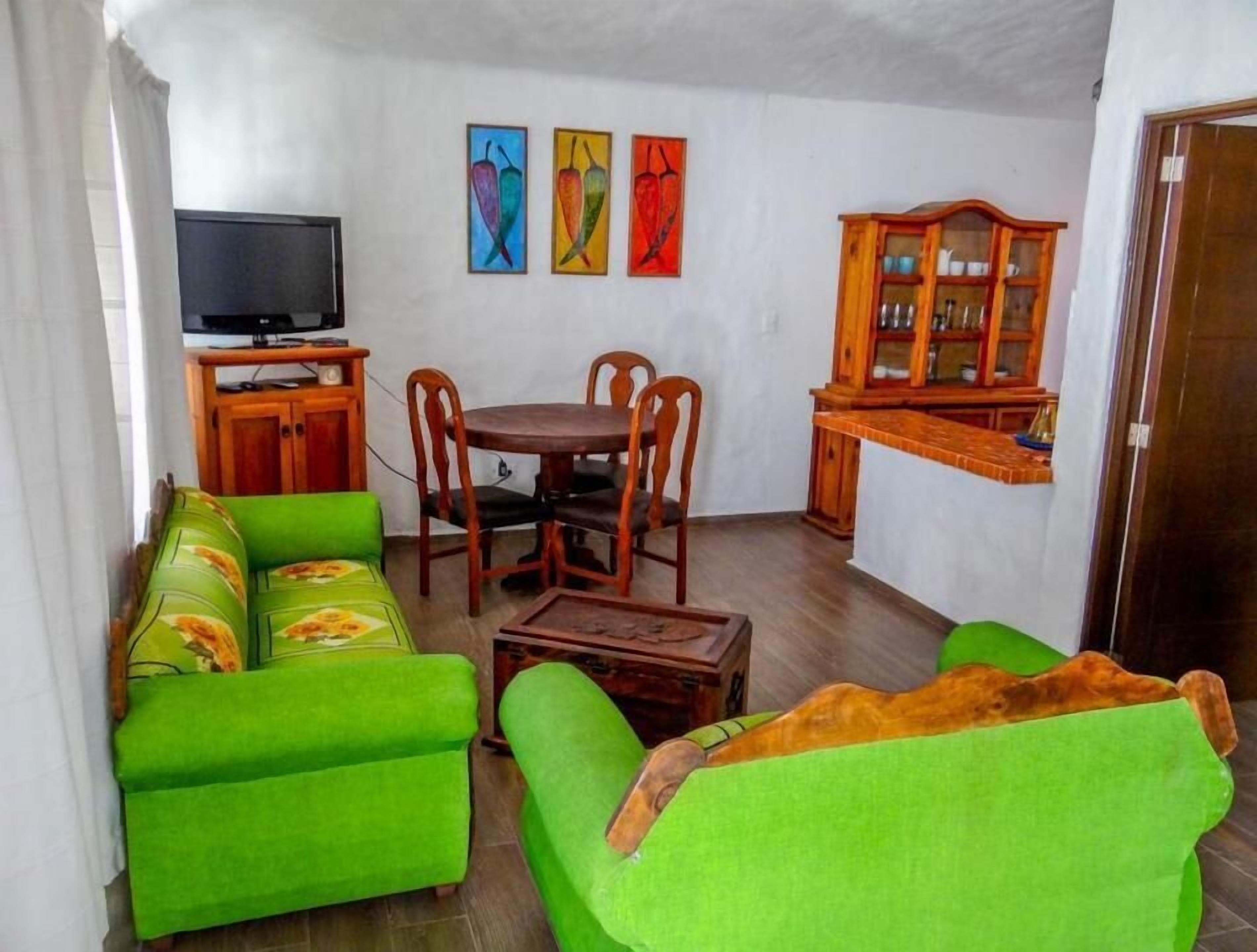 Ein Wohnzimmer mit grünen Sofas, einem Holzespeisetisch mit Stühlen, einem Fernseher auf einem Schrank und einem Schrank mit Glastüren.