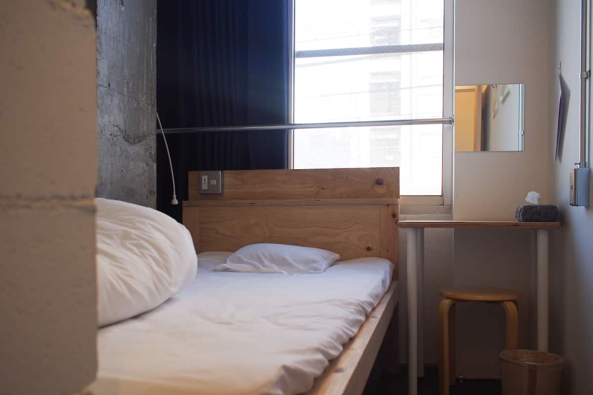 A single bed with white linens in a room with a wooden headboard, a small wooden stool, a trash bin, a mirror on the wall, and a window with curtains.