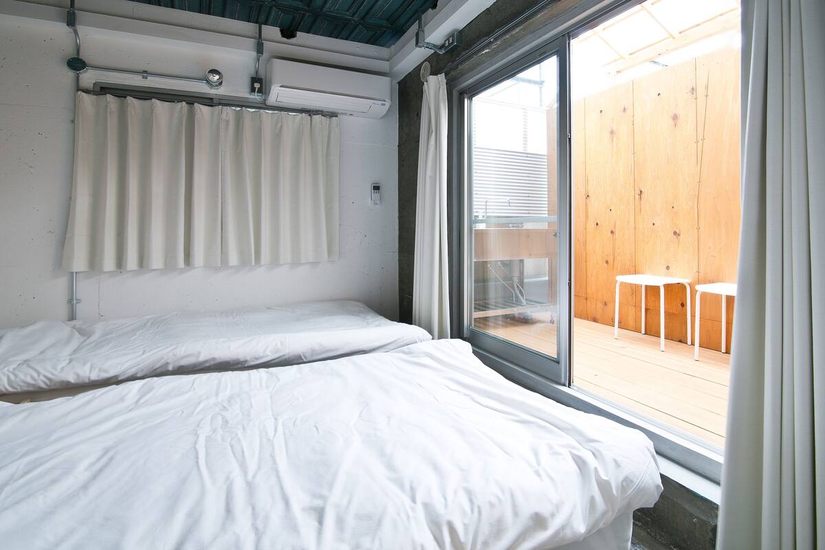 A room with two beds, a window with curtains, and a view of an outdoor wooden deck.